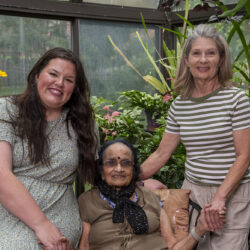 Three generations of women smile and pose together in a plant-filled greenhouse, with the elderly woman seated between two younger women.