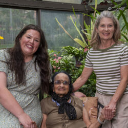 Three generations of women smiling together in a greenhouse with plants and flowers behind them.