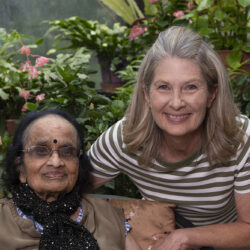 Two smiling women pose together in a lush garden with pink flowers, one seated wearing glasses and a scarf and the other leaning in from the right.