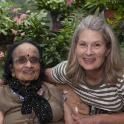 Two women posing together in a garden, smiling at the camera, with flowering plants in the background.