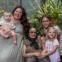 Grandmother sitting with her daughter, two granddaughters, and a baby in a greenhouse, all smiling.