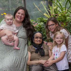 Multigenerational family portrait in a greenhouse: grandmother seated with mother holding a baby and two smiling girls beside them.