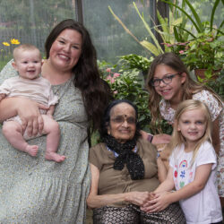 Family group in a greenhouse: grandmother seated center with two granddaughters beside her and a mother holding a baby on the left.