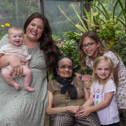 Grandmother seated center with two granddaughters and a mother holding a baby in a sunlit greenhouse, all smiling.