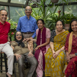 Six adults posing and smiling in a greenhouse surrounded by potted plants.