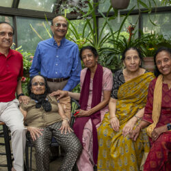 Group of seven people posing together in a glass greenhouse with lush potted plants around them, all smiling mostly seniors and adults.