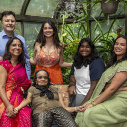 Group portrait of six Indian relatives in a greenhouse, smiling around an elderly woman seated center amid lush plants.