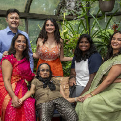 Group portrait of six women and an elderly woman gathered in a leafy greenhouse, smiling at the camera; colorful traditional outfits and plants surround them.
