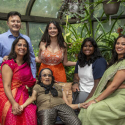 Multigenerational Indian family posing together in a glass greenhouse filled with plants, all smiling at the camera.
