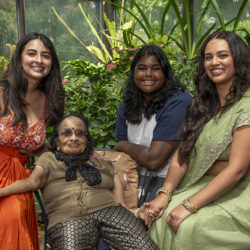 Four women of different generations smile and hold hands in a greenhouse, with the elderly woman seated center surrounded by plants.