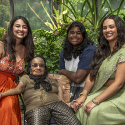Four women pose together in a greenhouse, smiling; an elderly woman sits center while three younger women sit and kneel beside her among potted plants.