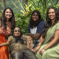 Four women of different ages sit together in a greenhouse, smiling and holding hands in a bright plant-filled setting