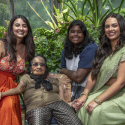 Four women of different generations smile together in a sunny greenhouse, with an elderly woman in a wheelchair at the center, holding hands with them.