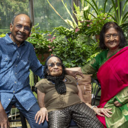 Three elderly adults smile together in a greenhouse surrounded by leafy plants.