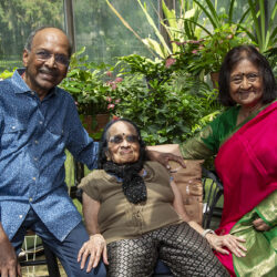 Three generations posing in a sunny garden: a man on the left, an elderly woman centered, and another woman in a pink saree on the right, all smiling.