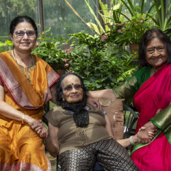 Three Indian women posing together in a sunlit garden, with the elderly woman seated between two smiling helpers holding her hands