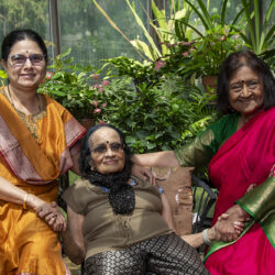 Three Indian women in colorful sarees smile and hold hands in a lush greenhouse garden.