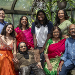 Multigenerational Indian family of nine posing together in a sunlit greenhouse with hanging plants and pots, smiling at the camera.