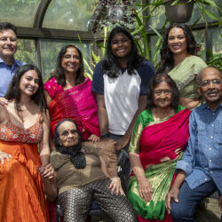 Family group portrait in a glass greenhouse, everyone smiling in colorful outfits.