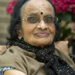 Portrait of an elderly Indian woman with gray hair and a black glittery scarf, wearing glasses and a bindi, seated outdoors with pink flowers behind her.