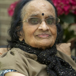 Elderly Indian woman wearing glasses and a black sequined scarf, smiling outdoors with pink flowers in the background.